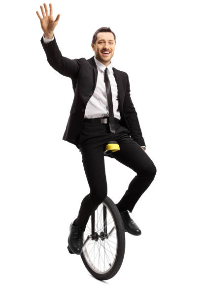 istock Young man in a suit riding a unicycle and waving at the camera 1158756423
