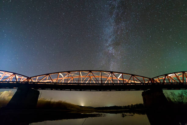 istock Illuminated metal bridge on concrete supports reflected in water on dark starry sky with Milky Way constellation background. Night photography concept. 1158757315