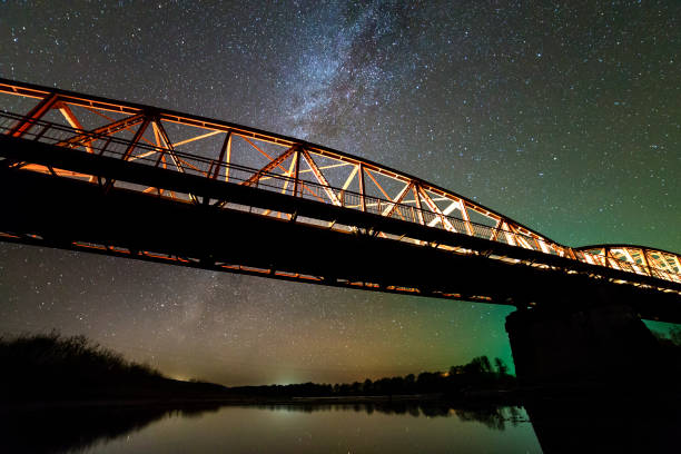 istock Illuminated metal bridge on concrete supports reflected in water on dark starry sky with Milky Way constellation background. Night photography concept. 1158757318