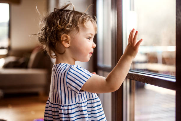 A toddler girl standing by window indoors at home, looking out. A happy toddler girl standing by window indoors at home, looking out. kid waves stock pictures, royalty-free photos & images