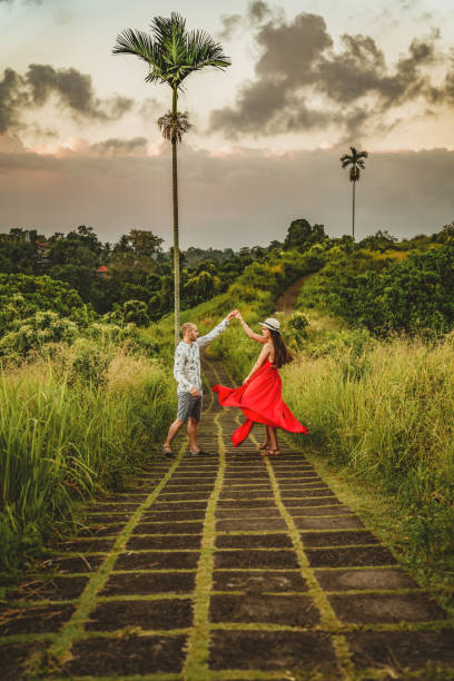 On an awesome outdoor adventure Side view of a Young beautiful woman and men dancing on Campuhan Ridgeway of artists, in Bali, Ubud. Beautiful calm sunny morning. Photo of a girl in a red dress and her boyfriend on the path of the artist. Bali trip. Tropics. Campuhan Ridge Walk. malay couple full body stock pictures, royalty-free photos & images