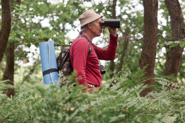 istock Profile of old traveller going camping alone, checking surroundings, looking through binoculars, fond of active lifestyle, taking all necessary equipment, wearing red sweatshirt, hat and backpack. 1158760962