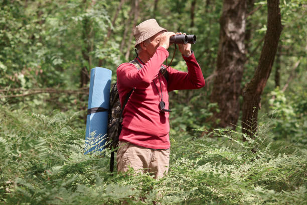 istock Profile of active hicker looking through binoculars, having compass on his neck, wearing sweatshirt and trousers, taking backpacker and and sleeping pad with him, going camping. Activities concept. 1158760969