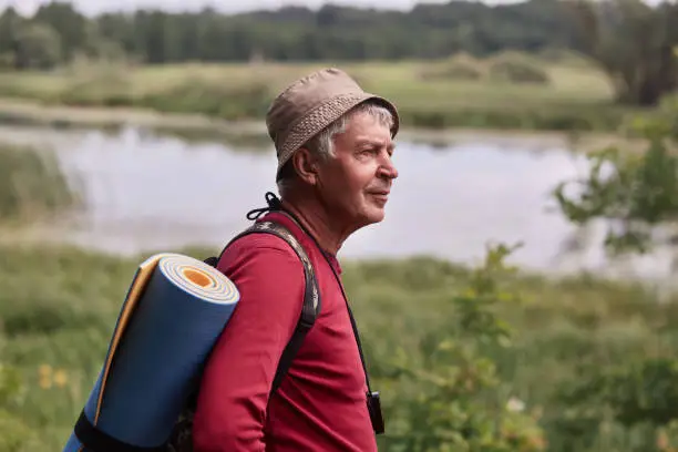 Profile shot of eldery man with backpack and blue rug, wearing red casual sweater and cap, enjoing beautifulnature near river and looking far away. Backpacking and active recreation concept.