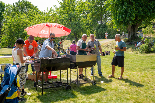 Jakobsberg, Sweden - June 6, 2019: Close-up summer view of two males barbecue grilling outdoors on a green lawn in a park with people and parasols on the Swedish national day in Jakobsberg Sweden June 6, 2019.