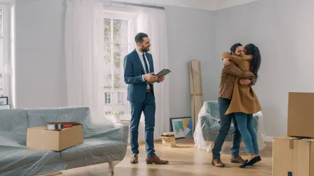 Photo of Real Estate Agent Shows Bright New Apartment to a Young Couple. Successful Young Couple Becoming Homeowners, Embraces and Hugs Each Other. Spacious Bright Home with Big Windows.
