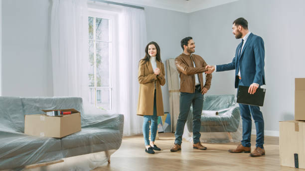 Professional Real Estate Agent Shows Bright New Apartment to Young Couple. Successful Young Couple Becoming Homeowners, Seal the Deal with Real Estate Broker by Handshake. Bright Home with Big Windows agente de bienes raíces profesional muestra nuevo apartamento brillante a la pareja joven. pareja joven exitosa convirtiéndose en propietarios de viviendas, sellar el trato con corredor de bienes raíces por apretón de manos. hogar brillante con grandes