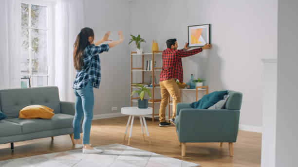 retrato de suspensão dos pares felizes na parede, o noivo move-lo, namorada diz-lhe quando o quadro está pendurado em linha reta. momento engraçado na vida dos pares novos. apartamento moderno e elegante. - home improvement fotos - fotografias e filmes do acervo