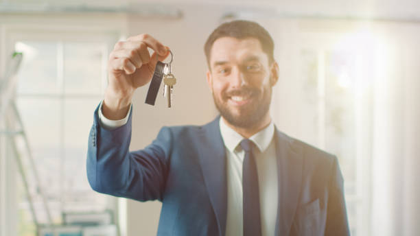 successful real estate agent in a suit smiles and offers keys from a new apartment. spacious new house for sale by professional real estate broker. shot with sun flare effect. - key real estate key ring house key imagens e fotografias de stock