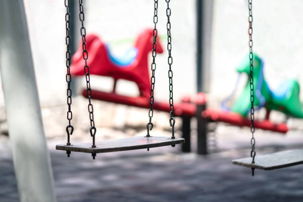 empty swing at a playground. sad dramatic mood for negative themes such as bullying at school, child abuse, pedophilia, traumatic childhood or kidnap. seesaw in the background. - swing playground empty abandoned imagens e fotografias de stock