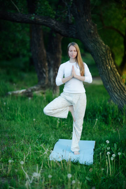 young woman in white clothes doing yoga, hands at the heart chakra in front of the chest namaste among trees at sunset, standing on the rug in  lush meadow stock photo