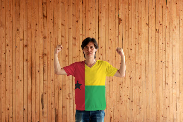 hombre con guinea bissau color de la bandera de la camisa y de pie con el puño levantado ambos en el fondo de la pared de madera. - guinea bissau flag fotografías e imágenes de stock