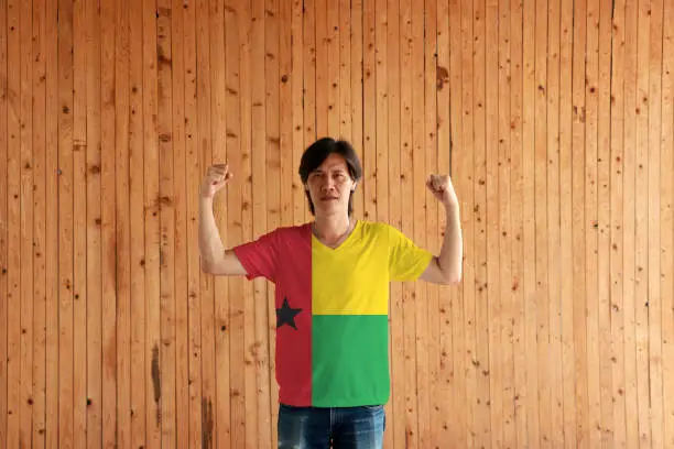 Photo of Man wearing Guinea Bissau flag color of shirt and standing with raised both fist on the wooden wall background.