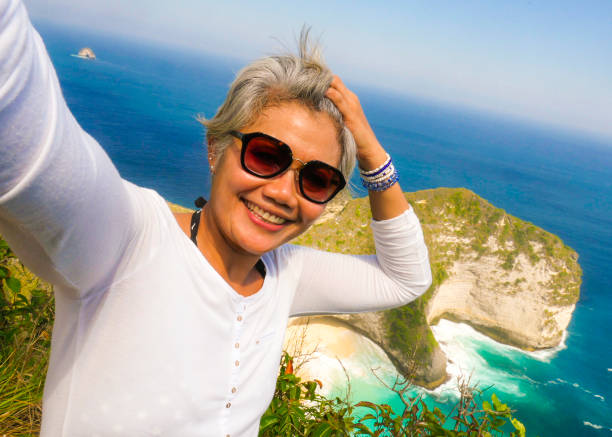 middle age 50s happy and cheerful Asian woman with grey hair taking selfie with mobile phone at beautiful tropical beach island smiling at cliff viewpoint enjoying Summer holidays travel destination stock photo