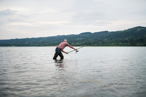 Senior fisherman in water casting with fishing rod in overcast weather condition.