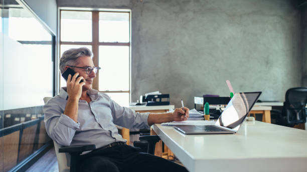 Businessman in office talking on phone Businessman on the phone sitting at the laptop in his office. Male business professional in office talking on cell phone. mature men photos stock pictures, royalty-free photos & images