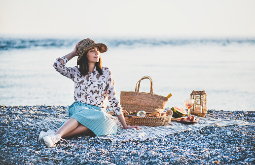Summer beach picnic at sunset. Young woman in hat sitting on blanket and having weekend picnic outdoor at seaside with fresh seasonal fruit and tray full of of tasty appetizers and looking away