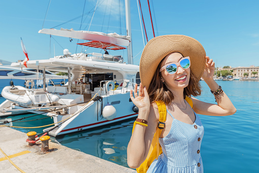 Young happy woman in hat with backpack standing near sea port with luxury yachts. Travel and vacation concept