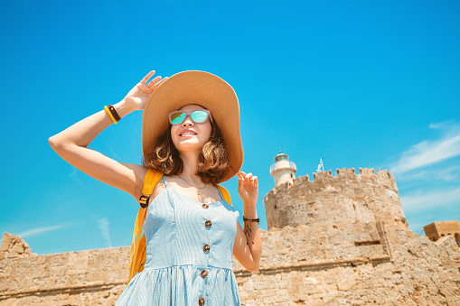 Happy tourist woman on vacation posing with hat and backpack in front of the old ruined fort and lighthouse in Rhodes, Greece