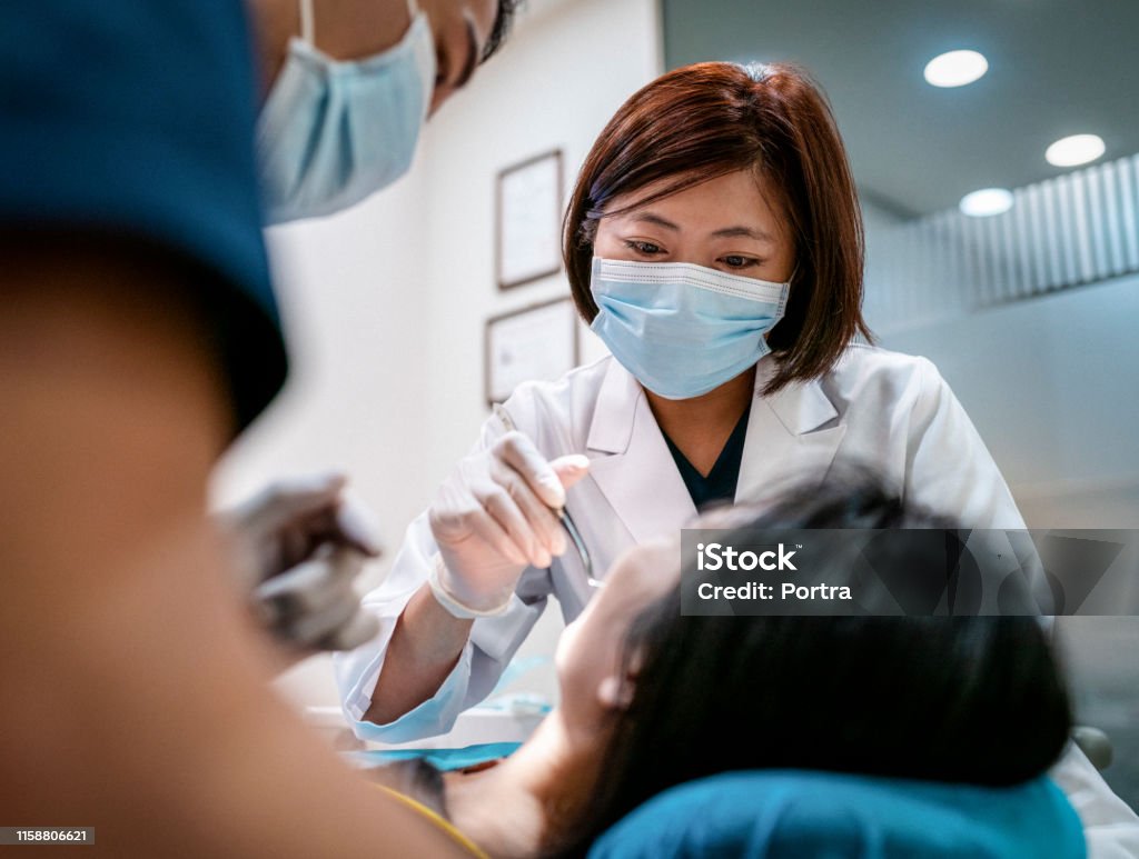 Dentist with male assistant treating female patient Female dentist wearing surgical mask treating woman. Male coworker is assisting mid adult healthcare worker. Medical professionals are examining patient in clinic. Dentist Stock Photo