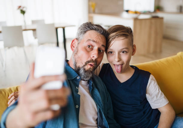 istock Father with small son sitting on sofa indoors, grimacing when taking selfie. 1158815057