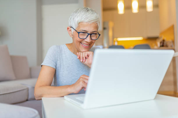 starsza kobieta pracująca na laptopie, uśmiechnięta. - biała kobieta czyta w okularach zdjęcia i obrazy z banku zdjęć