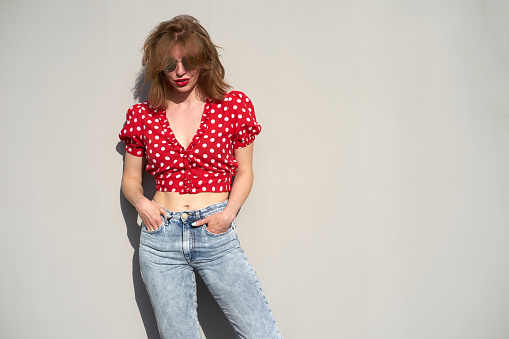 Redhead caucasian girl in red  polka-dot pattern blouse and jeans on grey background