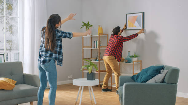 happy couple hanging picture on the wall, boyfriend moves it, girlfriend tells him when the frame is hanging straight. moment drôle dans la vie du jeune couple. appartement moderne élégant. - home decorating photos photos et images de collection