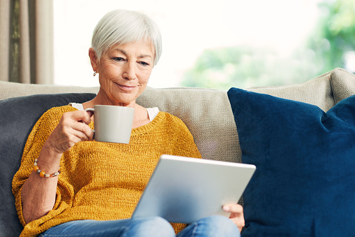 Shot of a senior woman using a digital tablet and having coffee on the sofa at home