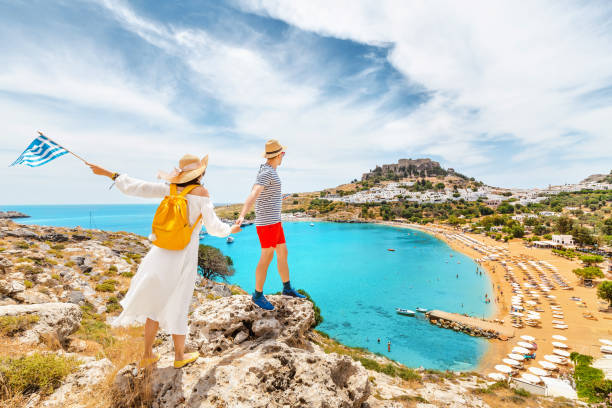 un couple d'amis amoureux du drapeau grec admirent la vue grandiose sur la baie de la mer et la plage près de l'ancienne ville de lindos. lune de miel et voyage - lindos photos et images de collection