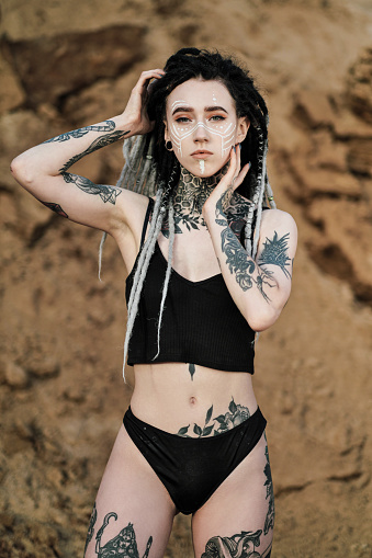 ✓ Beautiful sexy woman in black underwear. The body is covered with many  tattoos. Dreadlocks on the head . Posing against sandy career and black  fabric. Stock Photos