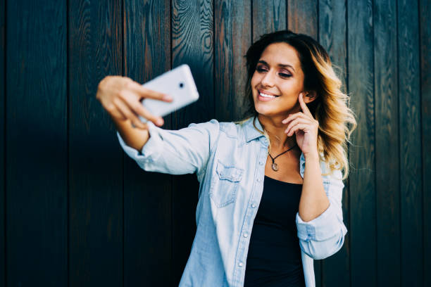 istock Cheerful female blogger dressed in stylish outfit posing while making selfie on cellular camera.Pretty positive student enjoying recreation during standing outdoors next to wall with copy space 1158822079