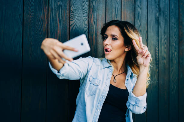 istock Young charming blogger posing for cellular camera while making selfie and showing peace.Cool female student fooling during taking photo and putting tongue on lips standing near to advertising area 1158822084