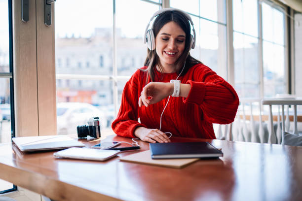 Cheerful hipster girl listening new bestseller audiobook using application and earphones sitting at coffee shop, positive teenager enjoying funny playlist and checking time on wearable smartwatch chica hipster alegre escuchando nuevo libro de audio bestseller utilizando la aplicación y auriculares sentados en la cafetería, adolescente positivo disfrutando de lista de reproducción divertida y comprobar el tiempo en reloj inteligente usable