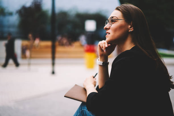 istock Young charming brunette woman in cool eyeglasses for better vision spending leisure time at street and planning working schedule.Cute hipster girl looking away while sitting at street and thinking 1158824265