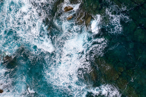 Sea waves seen from above, while splashing and flowing through dangerous rocks. Blue and cold hues. Aerial view.