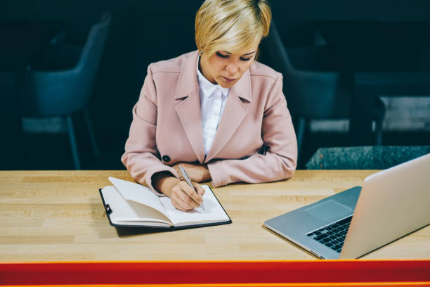 istock Pensive mature female entrepreneur writing down plan in notepad working at laptop computer in office.Businesswoman 50 years old noting information in notebook sitting at wooden table with netbook 1158829548