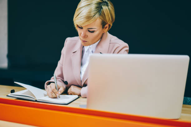 istock Pensive mature female professor preparing training seminar writing down notes of organization process in notepad working at modern netbook.Businesswoman 50 years old noting financial schedule 1158829555