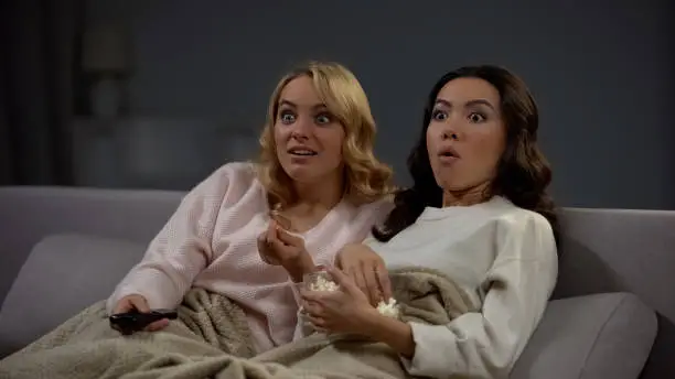 Photo of Two shocked women eating popcorn and watching interesting TV program, rest