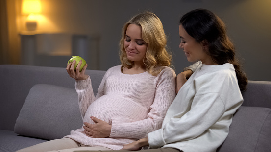 Pregnant woman holding apple talking with friend about diet for baby health