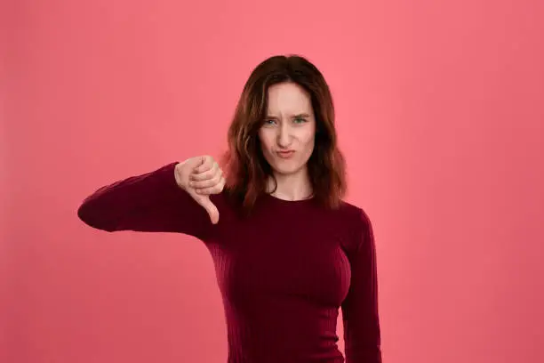 Unhappy young lady standing isolated over dark pink background with a gesture of thumb down symboling negative appraisal.