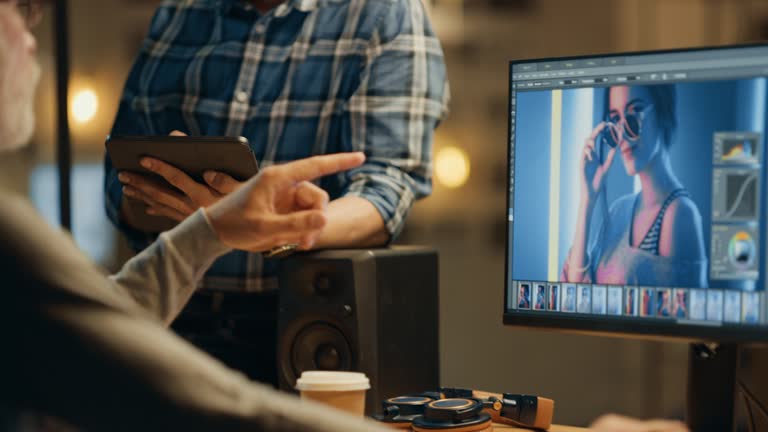 In the Evening Creative Middle Aged Photo Editor Works on a Desktop Computer Uses Image Editing Software, Has Conversation with Younger Specialist who Uses Tablet Computer. Stylish Office in the Evening