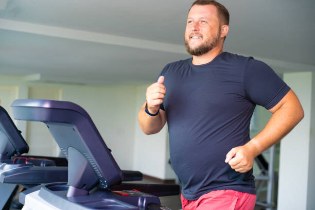 smiling full male runs on a treadmill in a gym. concept of weight loss and sport. side view smiling full male runs on a treadmill in a gym. concept of weight loss and sport. side view treadmill stock pictures, royalty-free photos & images