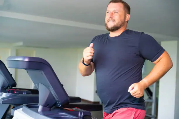 Photo of smiling full male runs on a treadmill in a gym. concept of weight loss and sport. side view