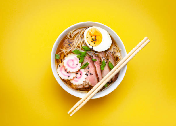 Tasty Japanese noodle soup ramen in white ceramic bowl with meat broth, sliced pork, narutomaki, egg with yolk on pastel yellow background. Traditional dish of Japan, top view, close-up, concept