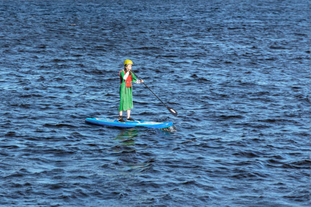 St. Petersburg, Russia - June 23, 2019: slim girl in a yellow helmet and a long green dress floating on the river on the SUP board