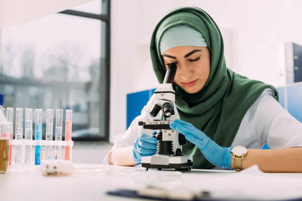 schöne muslimische wissenschaftlerin, die während des experiments im chemielabor durch das mikroskop schaut - religiöse kleidung stock-fotos und bilder