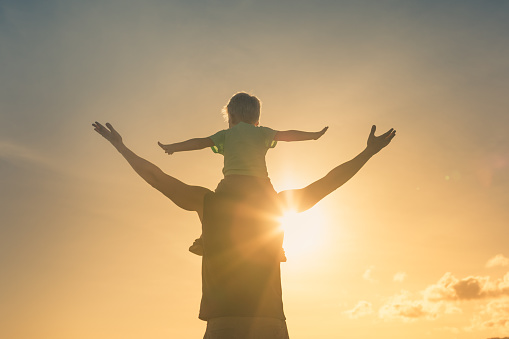 Happy father and son with arms outstretched playing together against sunset sky.