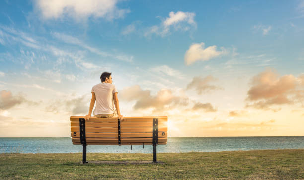istock Young thoughtful man sitting on park bench looking at ocean view 1158891141
