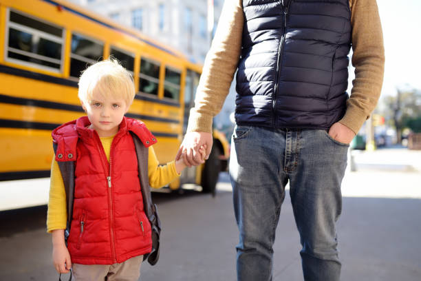 istock Elementary student hold hands his father near yellow school bus on background. 1158899679